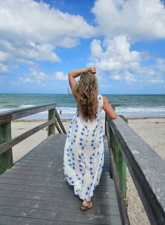 A woman in resort casual attire dress, it is a flowy white sleeveless tunic dress in white with blue flowers. The woman is standing on a boardwalk to the ocean.