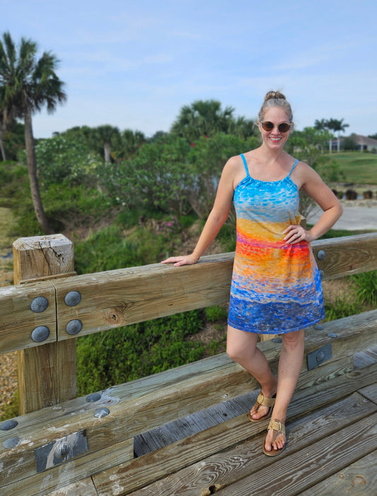 A woman in a cotton dress in a tropical location for vacation. She is standing on a bridge and her dress is knee length with a brigh sunrise pattern that is abstract.