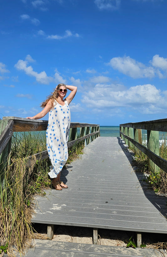 A woman wearing a flowing maxi rayon beach and vacation dress. The dress is sleeveless tunic style that is white with blue flowers. She is standing on a boardwalk to the beach and ocean. She is facing the camera sideways and has an arm up by her hair.