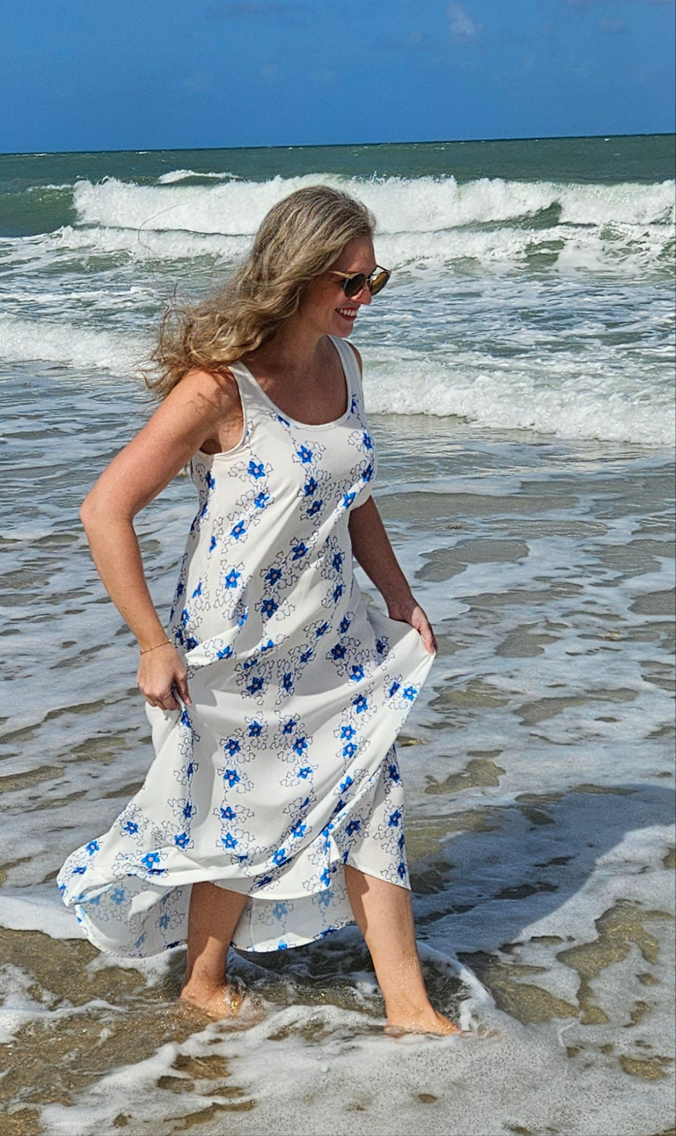 A woman on the beach in a beach vacation dress for a tropical vacation. It is a white rayon maxi dress with small blue flowers. She is walking in the ocean water, her dress flowing in the wind as is her hair.