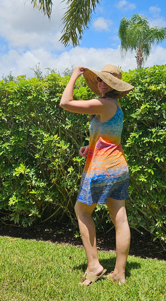 A woman wearing a cotton sundress with bright colors and knee length. The design is an abstract sunrise. She has a straw hat on and is standing in front of palm trees in a tropical location.