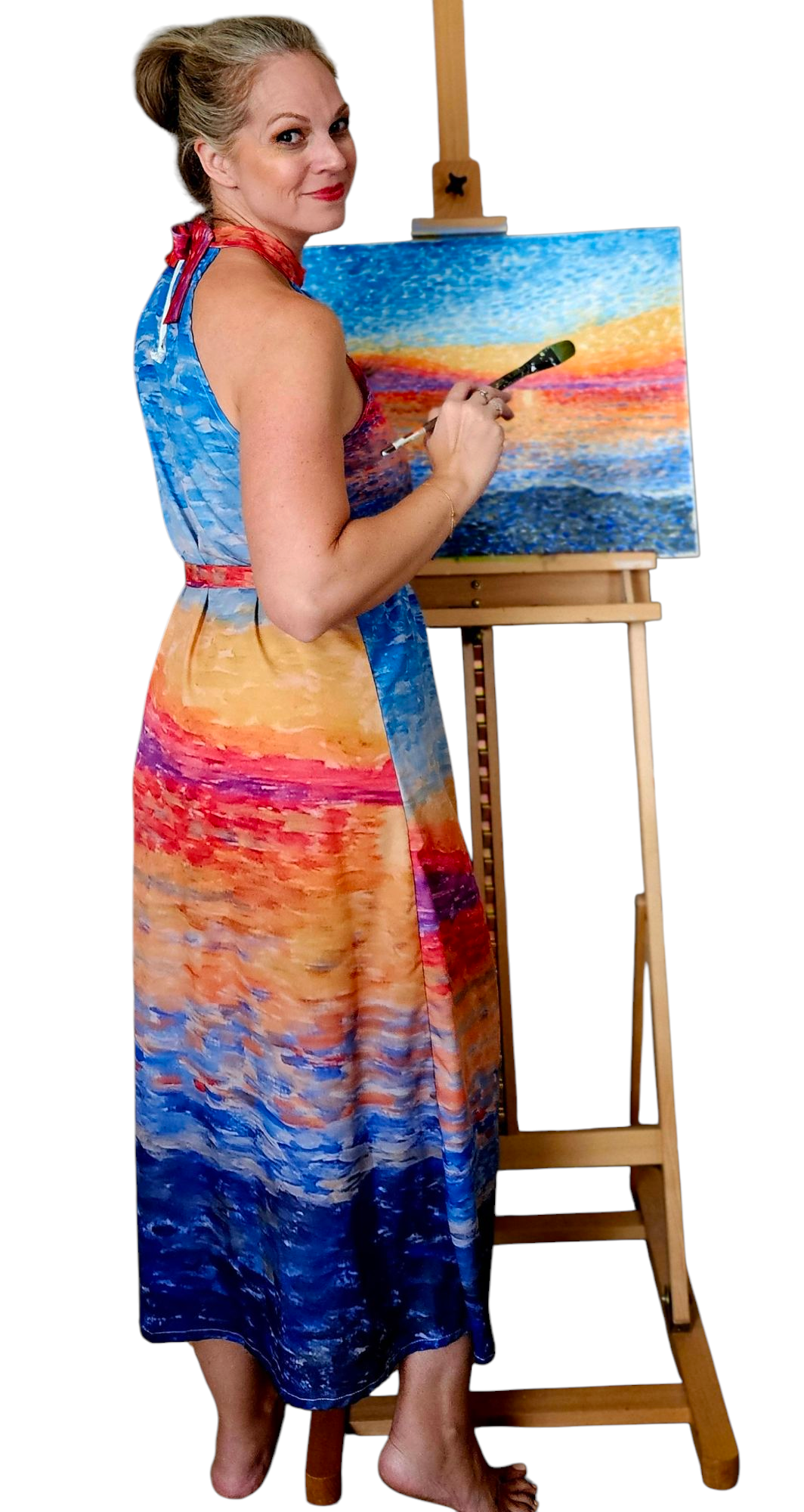 A woman is standing in front of a painting with a paintbrush. She is looking over her shoulder at the camera. She is wearing a halter resort and vacation dress in bright colors matching the sunrise painting in front of her. The dress has a high neck, tie waist, and is midi length.