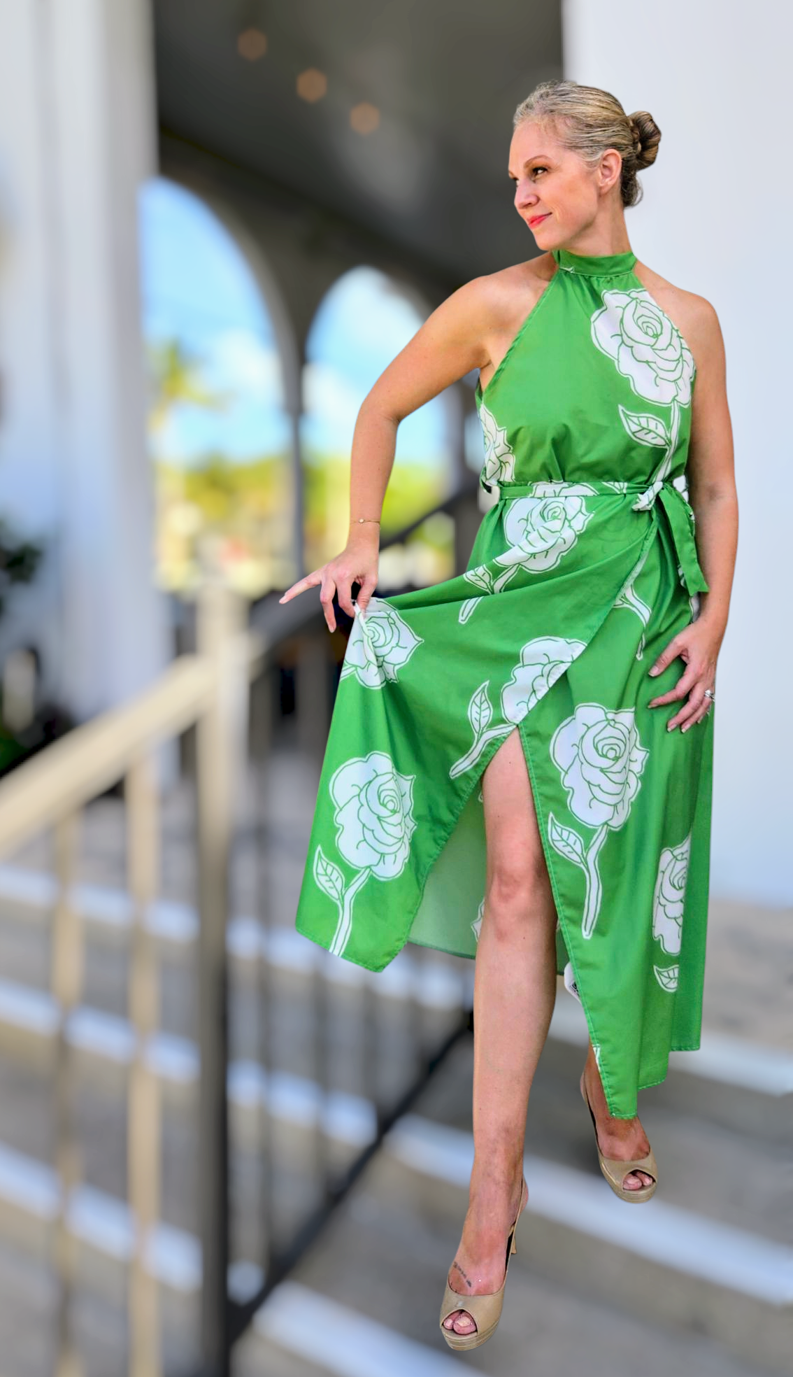 Woman in a green midi halter vacation and resort wear dress. The dress has a high neck with a tie waist and side slit, it is a wrap dress. The halter dress is a bright green with white roses as the design. 