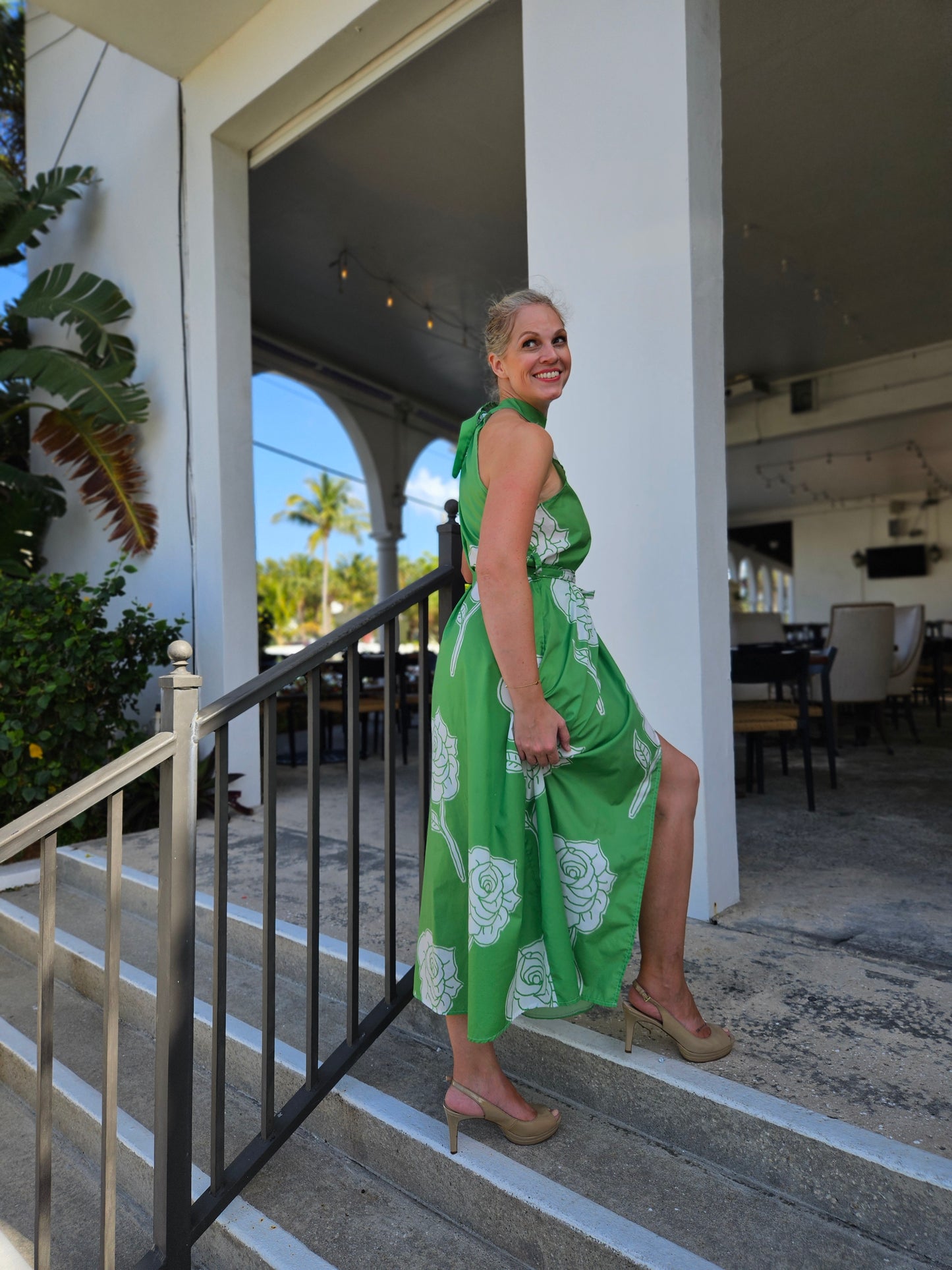 A woman wearing a green high neck, tie waist, side slit midi halter dress. She is walking up stairs into a restaurant. This dress is from the vacation and resort wear collection. 