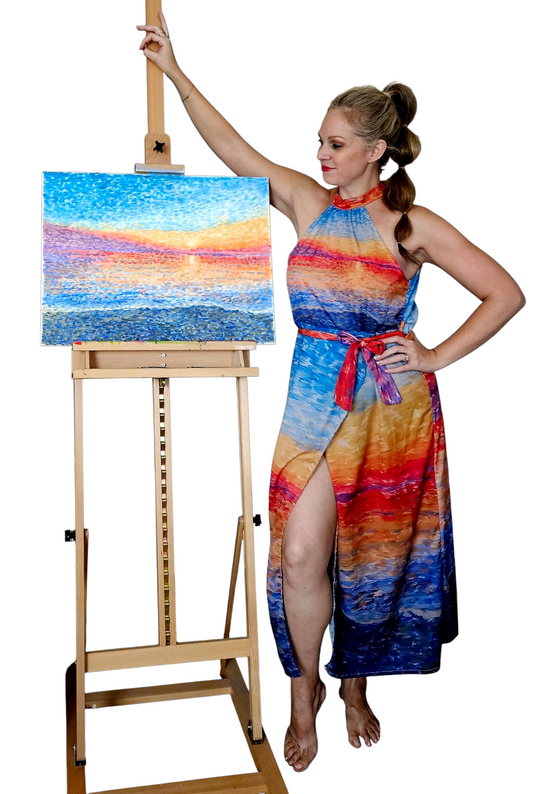 A woman wearing a resort halter vacation dress in bright colors. It is a high neck, tie waist, midi halter dress. It is a tropical vacation dress, and it matches a painting next to the woman.