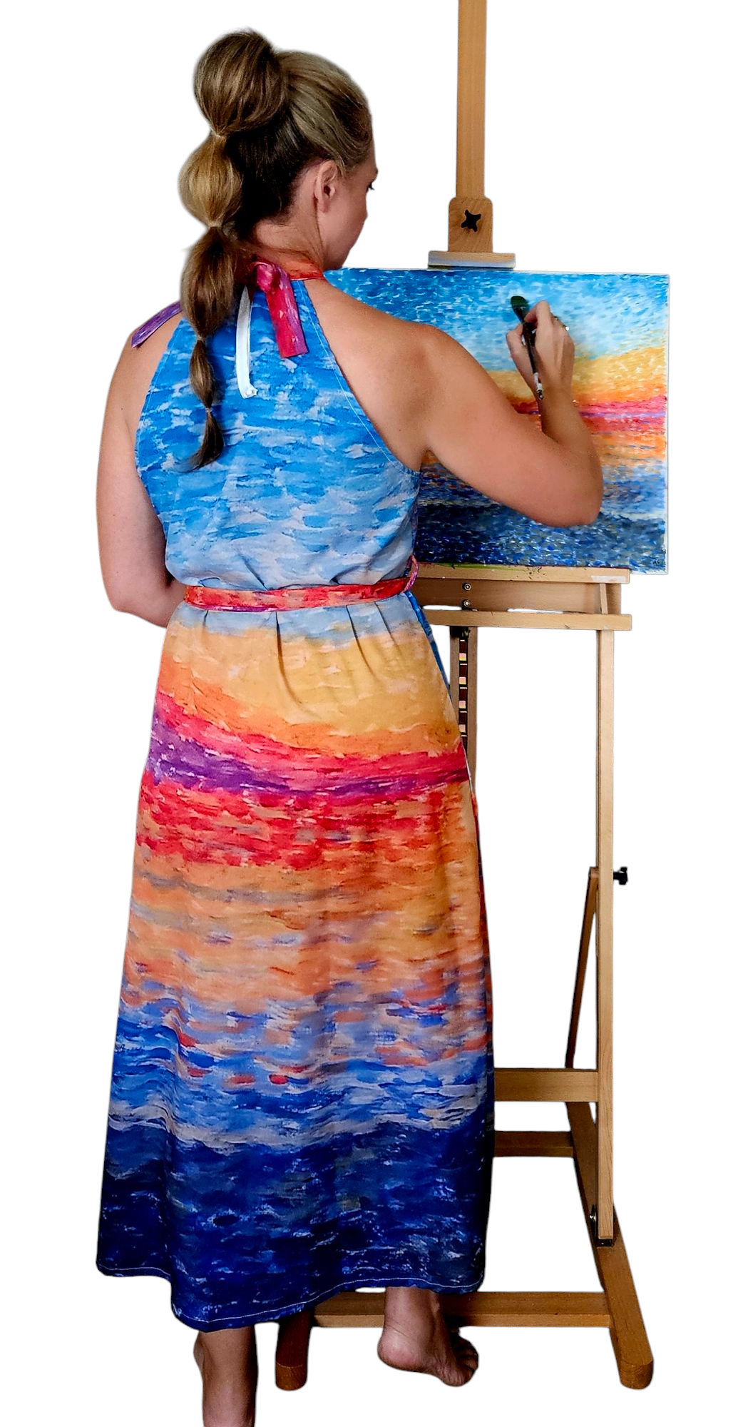 A woman in a halter vacation dress. The dress is a resort dress in bright colors matching the painting in front of her. The dress is high neck, tie waist, and midi length. She is holding a paintbrush.