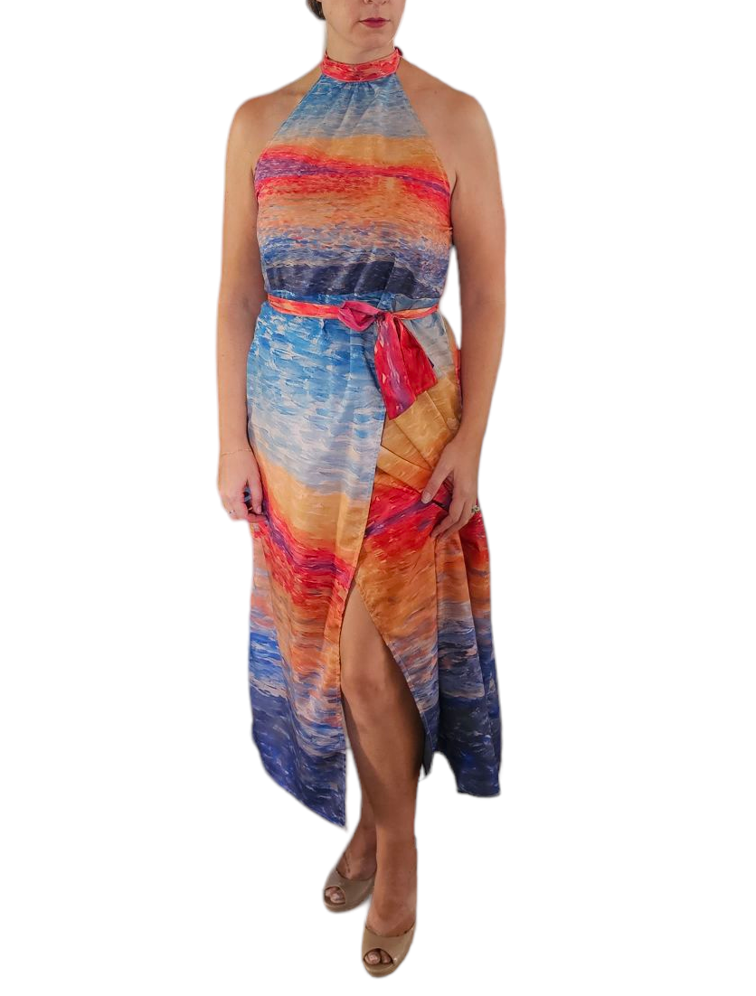 A woman wearing a high neck, tie waist, midi halter vacation dress. The dress has vibrant colors that are paint brush strokes of a beautiful sunrise.